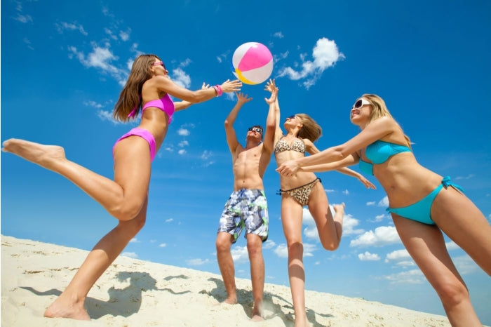 Four friends in swimsuits toss a beach ball under a vivid blue summer sky on a sandy beach.
