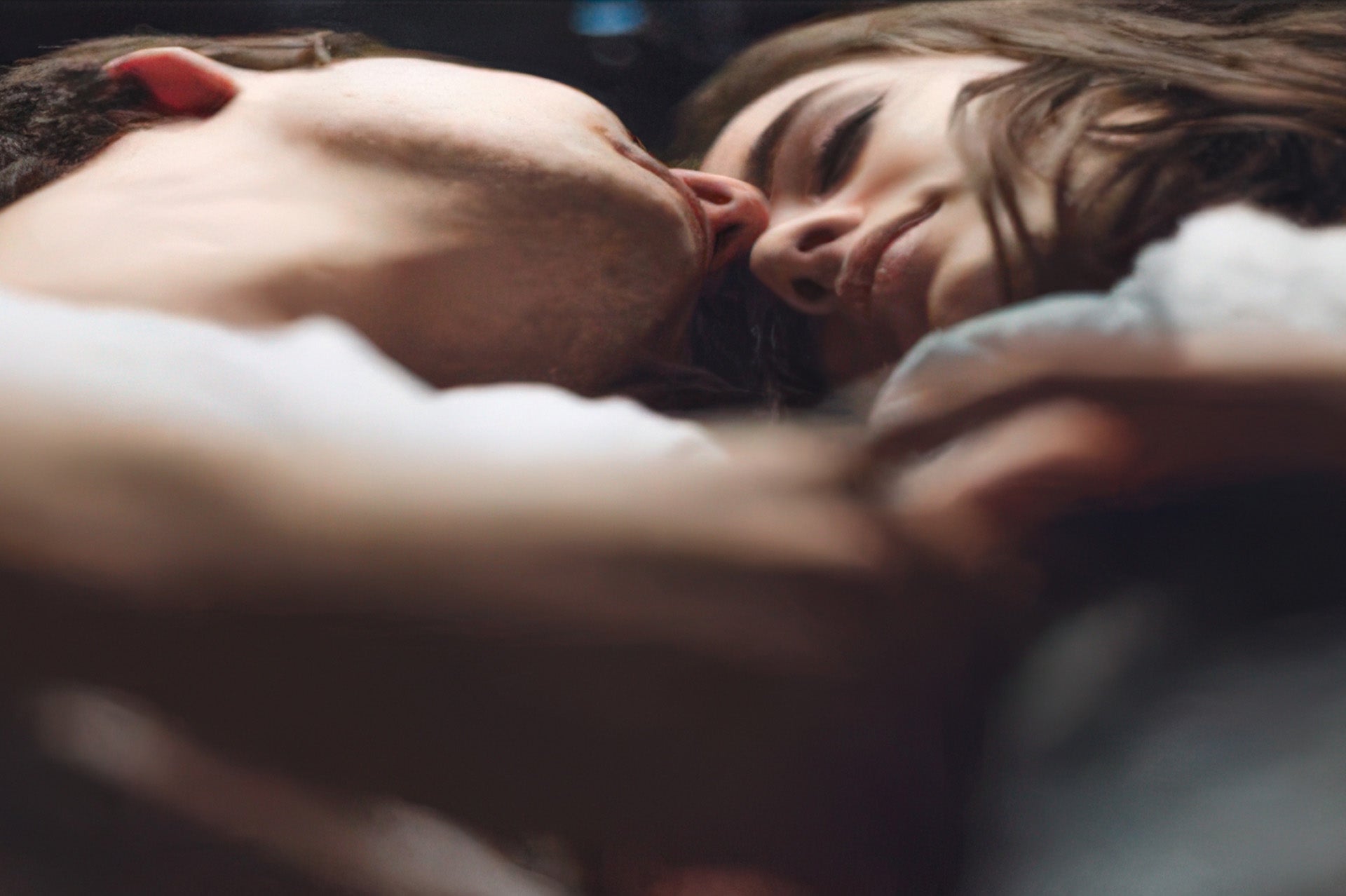 A couple lies close together on dark bedding, faces nearly touching in soft intimate lighting.