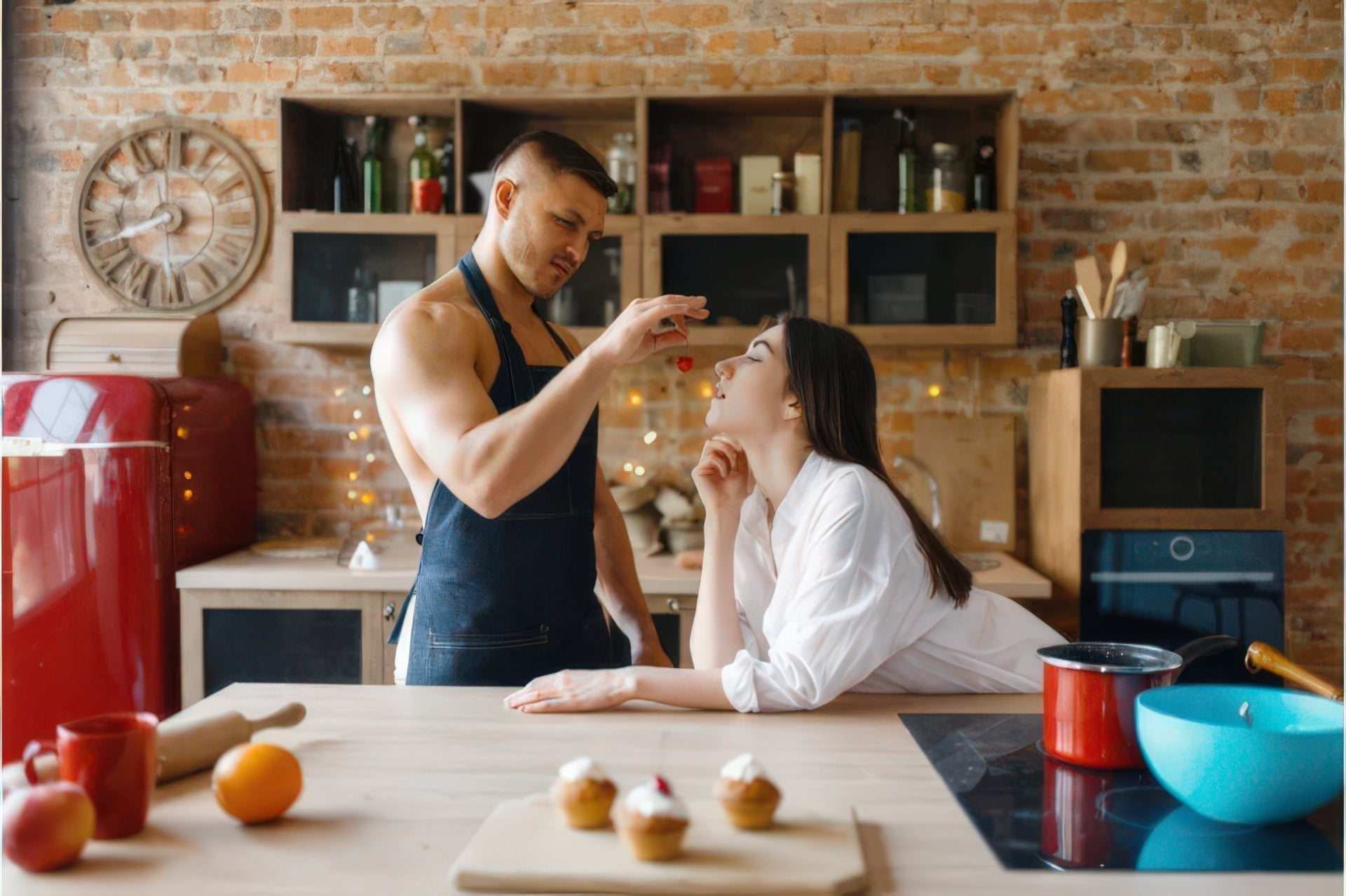 A muscular man in an apron playfully feeds a cherry to a smiling woman in a rustic kitchen.