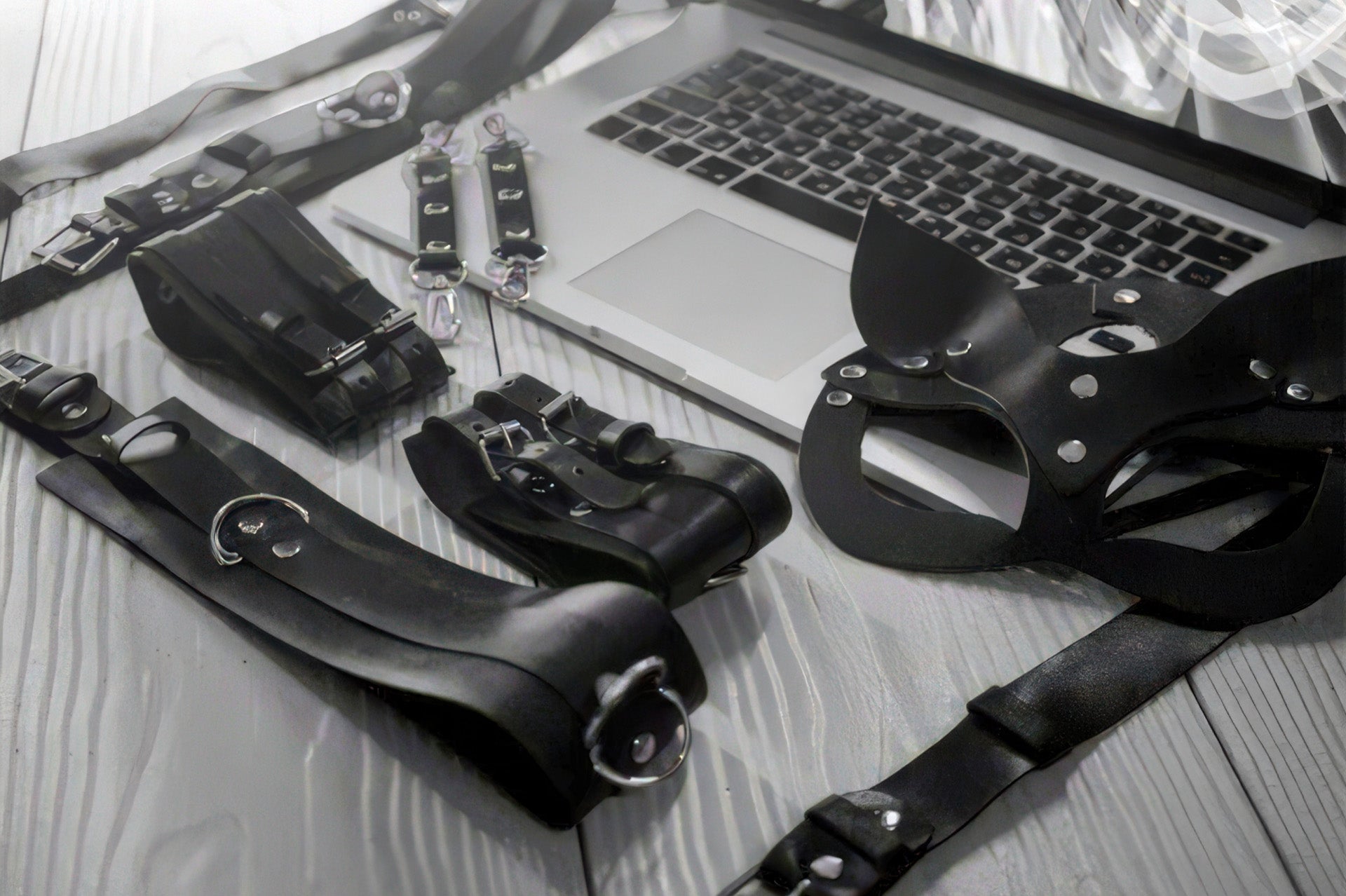 Black-and-white flatlay of leather BDSM accessories — cuffs, mask, and straps — spread beside a laptop.