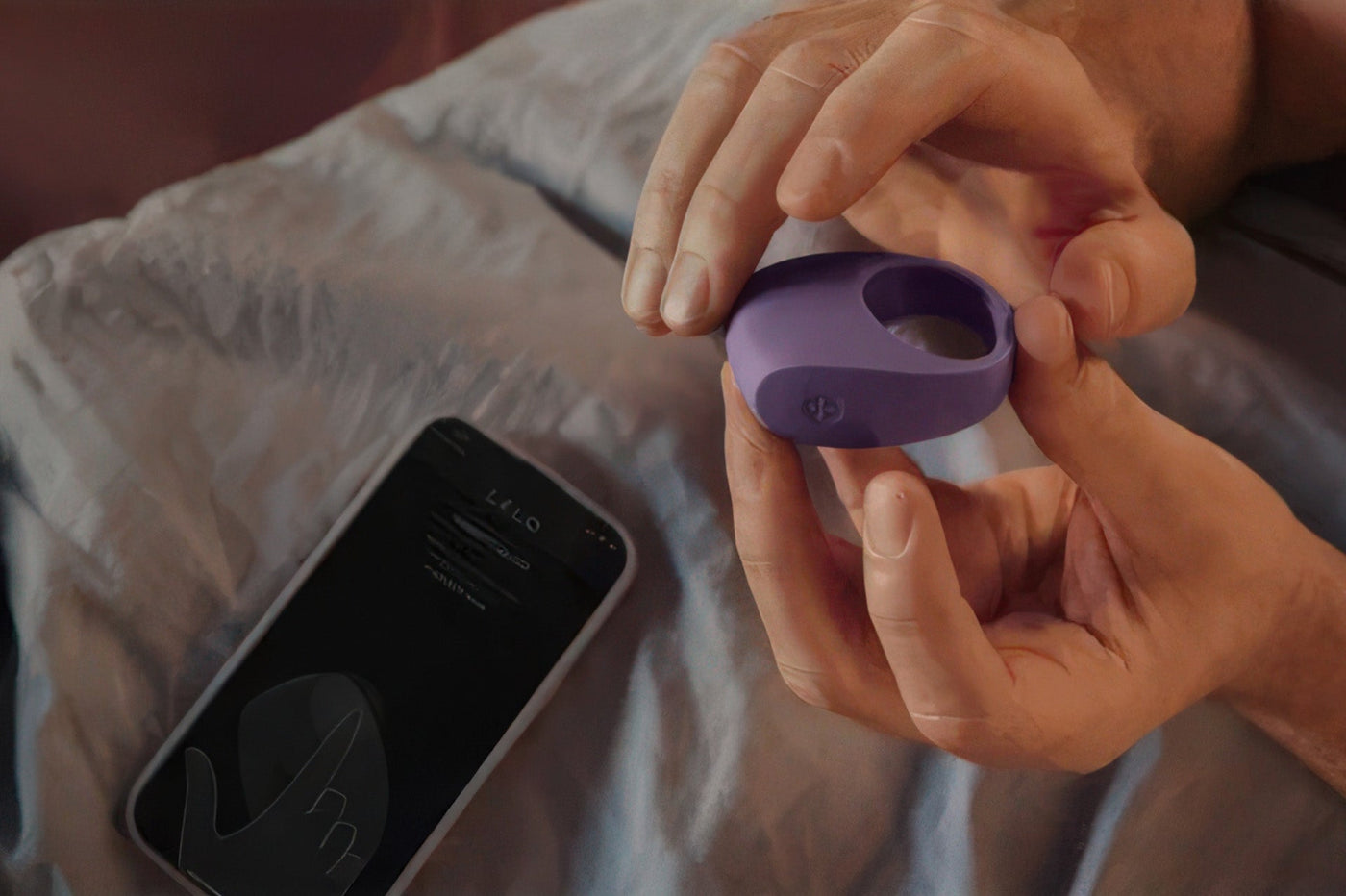 Hands hold a purple LELO vibrating couples ring over grey satin sheets next to a smartphone app.