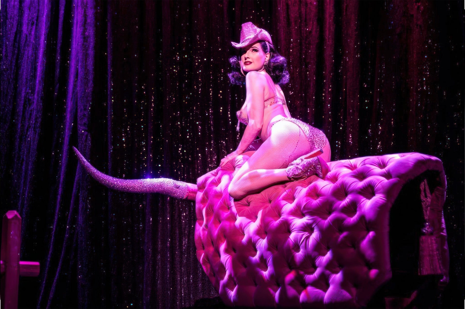 A burlesque dancer in a bejeweled costume perches on an oversized pink tufted saddle prop on a glittering stage.