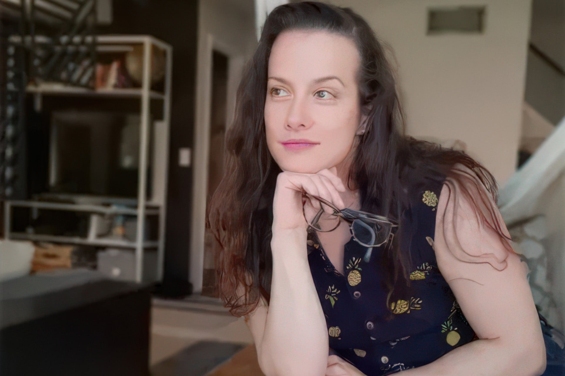 A dark-haired woman, Kelly Cupo, in a floral top sits pensively holding glasses in a warmly lit home interior.