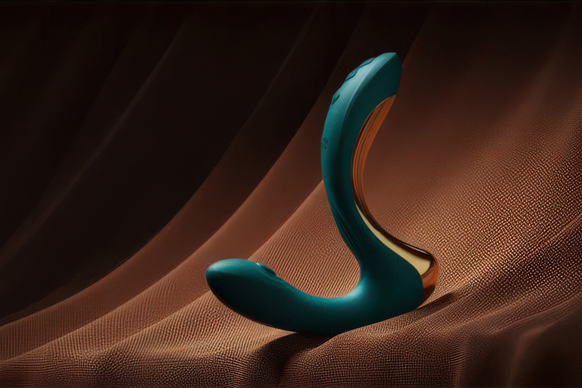 A teal and gold curved vibrator rests against rich brown draped fabric in dramatic studio lighting.