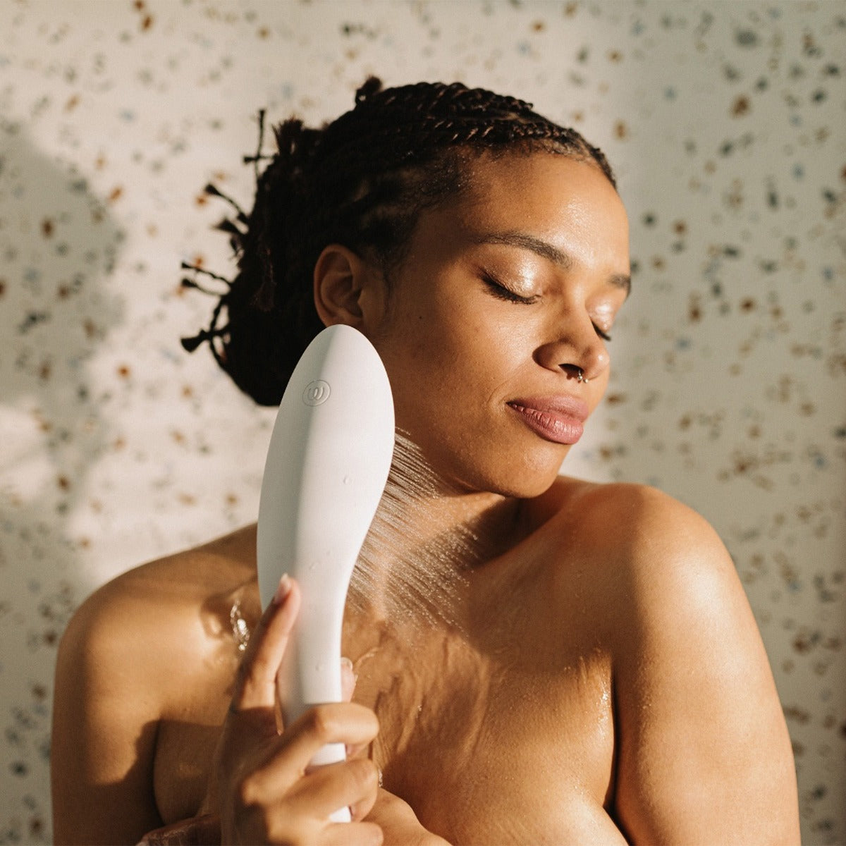 Woman spraying herself with a showerhead against a floral-patterned wall.