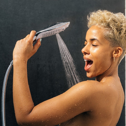 Woman holding a shower head with water spraying towards her face against a dark background