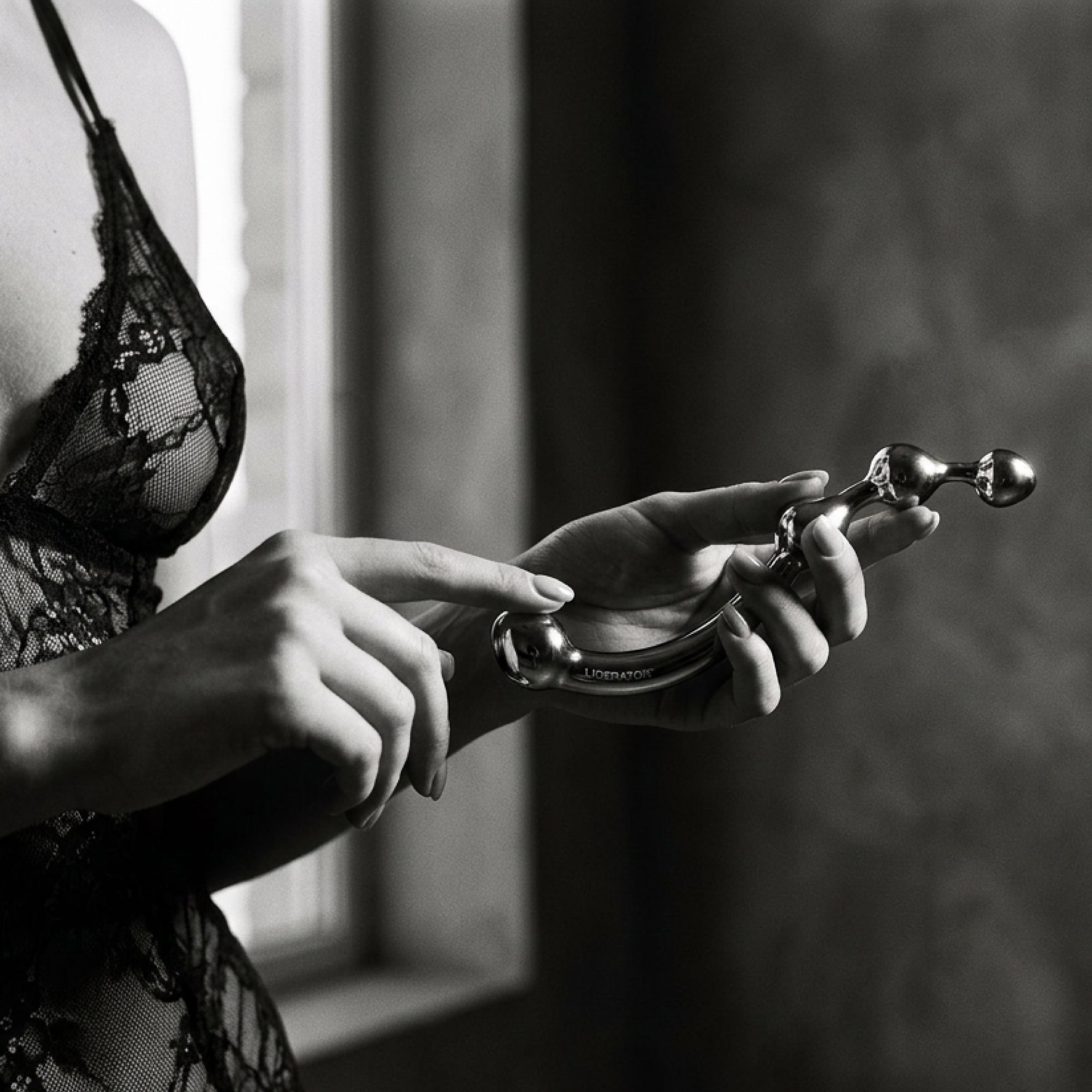 A black and white close-up of a person in sheer lace lingerie holding a curved metallic personal massager with a spherical head.