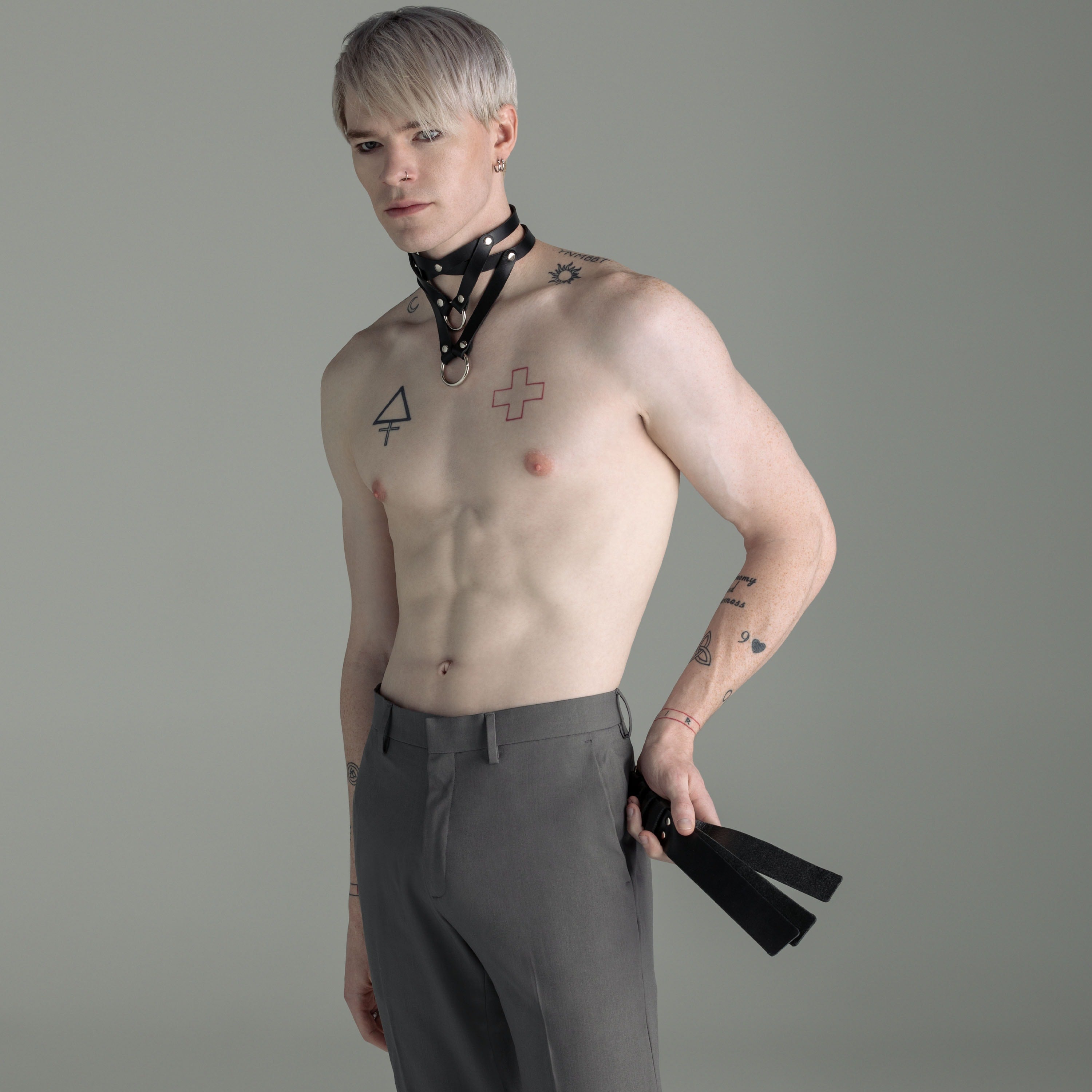 Man with tattoos wearing gray pants and a choker, holding a black leather slapper against a gray background