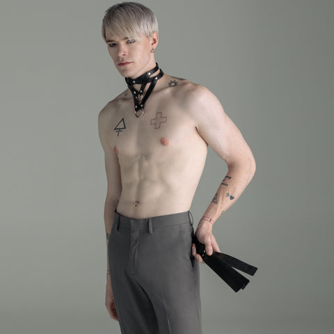 Man with tattoos wearing gray pants and a choker, holding a black leather slapper against a gray background