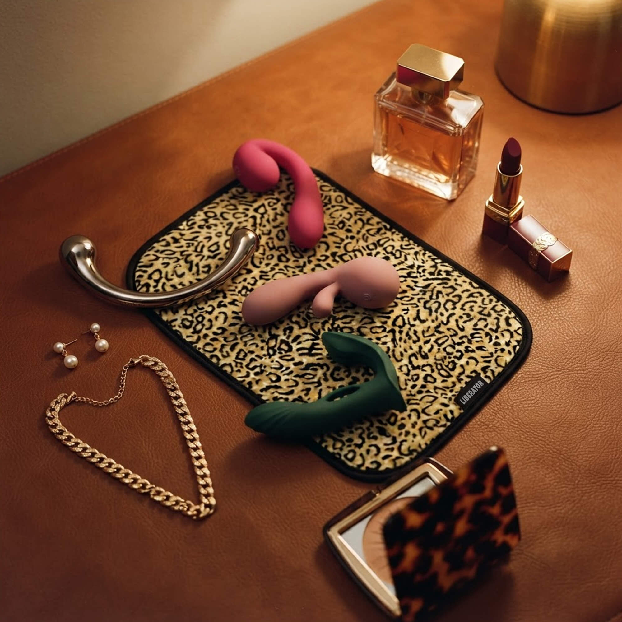 A styled flat-lay of intimate wellness products on a brown leather-like surface, including a pink curved wand massager, a pink dual-stimulation vibrator, a green curved prostate massager, a silver curved metal wand, a leopard-print toy pad, and other cosmetics and jewelry.