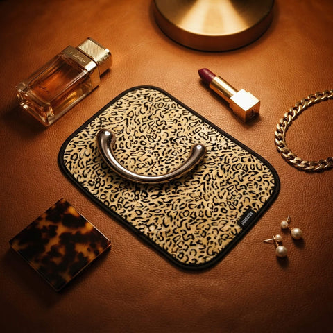A leopard-print toy pad with a silver metal wand massager, styled on a brown leather surface with luxury accessories including a perfume bottle, lipstick, jewelry, and a tortoiseshell case.