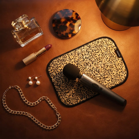 A black handheld wand massager on a leopard-print toy pad, styled on a warm brown leather-like surface with perfume, lipstick, earrings, pearls, and a mirror around it.