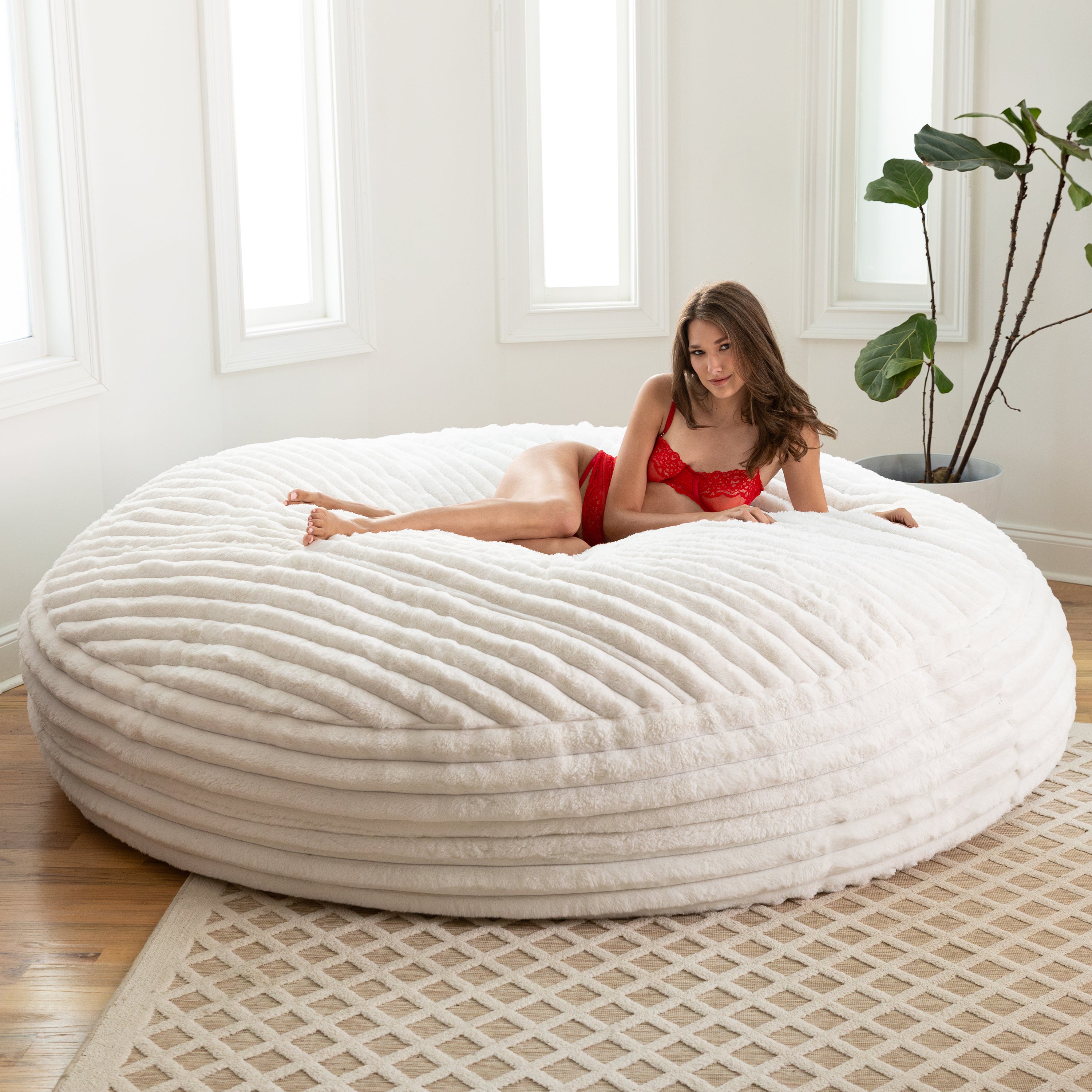 Woman in a red outfit lying on a large white textured bean bag chair in a bright room with a plant.