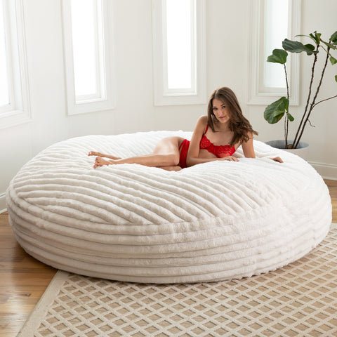 Woman in a red outfit lying on a large white textured bean bag chair in a bright room with a plant.